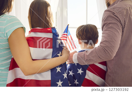Back view of a young family standing at window wrapped in us flag celebrating Independence Day. Back view of a young family standing at window wrapped in us flag celebrating Independence Day. 127303192