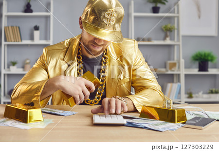Funny man in golden suit holding gold bar and counting profit with cash dollar money at table 127303229