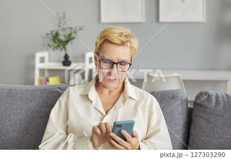 Senior mature woman using smartphone, browsing apps, sitting at home, looking busy at phone screen 127303290