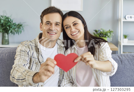 Young cheerful couple holding paper heart in hands sitting on sofa in living room at home. Young cheerful couple holding paper heart in hands sitting on sofa in living room at home. 127303295