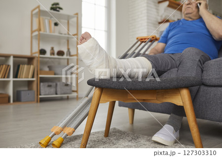 Elderly Man With Broken Leg Resting On Sofa At Home While Talking On The Phone 127303310