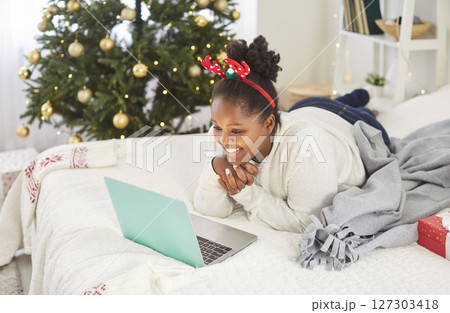 Girl lying on bed at home with Christmas tree using laptop for shopping or having online video call Girl lying on bed at home with Christmas tree using laptop for shopping or having online video call 127303418