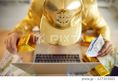 Top view of funny man sitting at desk with laptop, holding gold bar and dollars banknotes Top view of funny man sitting at desk with laptop, holding gold bar and dollars banknotes 127303881