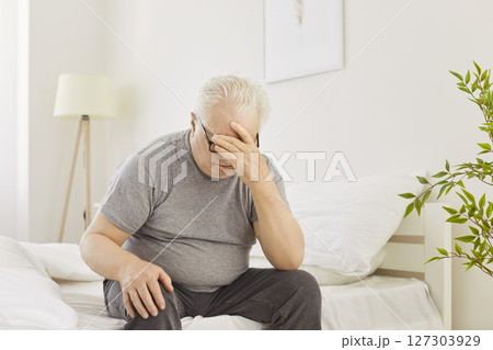 Sad depressed senior man with sleep problems and headache wakes up tired and sits on bed 127303929