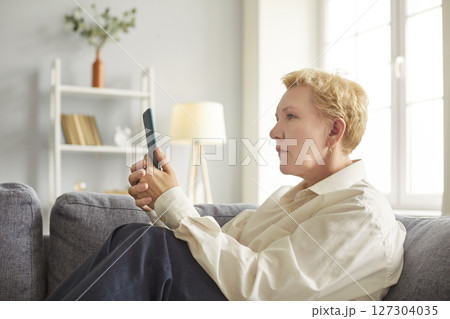 Senior concentrated older lady on couch at home, using applications on smartphone screen Senior concentrated older lady on couch at home, using applications on smartphone screen 127304035