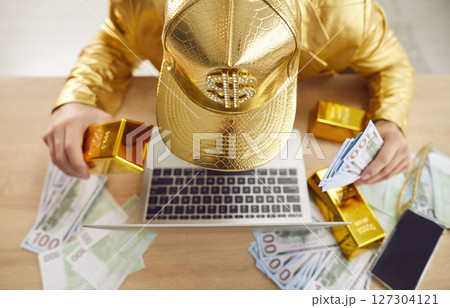Top view of young funny man sitting at desk with laptop, holding gold bar and dollars banknotes Top view of young funny man sitting at desk with laptop, holding gold bar and dollars banknotes 127304121