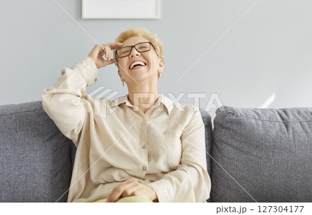 Portrait of happy laughing mature woman wearing glasses sitting on comfortable sofa at home Portrait of happy laughing mature woman wearing glasses sitting on comfortable sofa at home 127304177