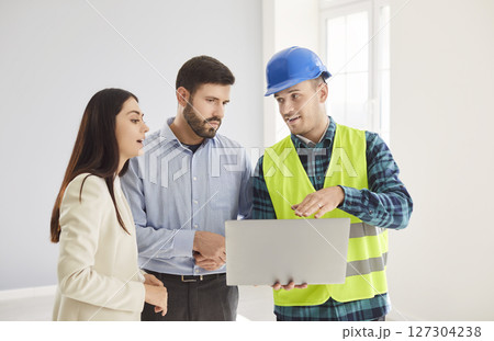 Construction worker in blue helmet showing apartment renovation plan on laptop to young couple. Construction worker in blue helmet showing apartment renovation plan on laptop to young couple. 127304238