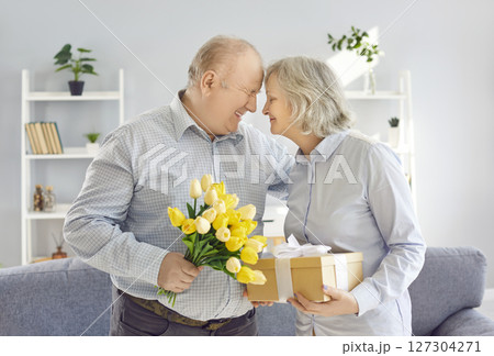 Happy senior couple standing at home with flowers bouquet and present gift box. 127304271