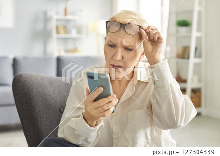 Shocked in worry, concerned mature woman in glasses holding smartphone, staring at phone screen 127304339