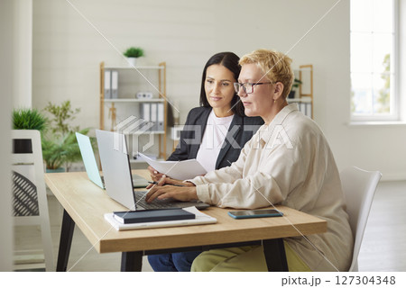 Two female colleagues working together at desk in office, using laptop and analyzing business data. Two female colleagues working together at desk in office, using laptop and analyzing business data. 127304348