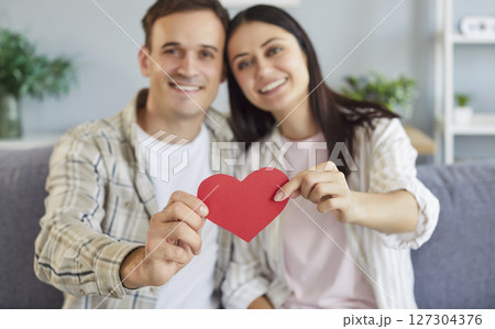 Young cheerful smiling brunette woman and man holding paper heart in hands sitting on sofa at home Young cheerful smiling brunette woman and man holding paper heart in hands sitting on sofa at home 127304376