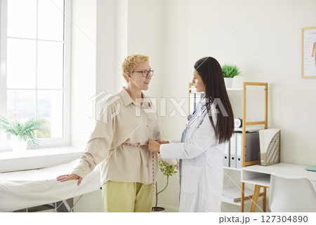Young woman doctor measuring waist of female patient with tape measure during examination in clinic. Young woman doctor measuring waist of female patient with tape measure during examination in clinic. 127304890