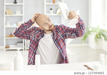 Young man with alopecia is holding a table mirror and looking at his balding head Young man with alopecia is holding a table mirror and looking at his balding head 127304891