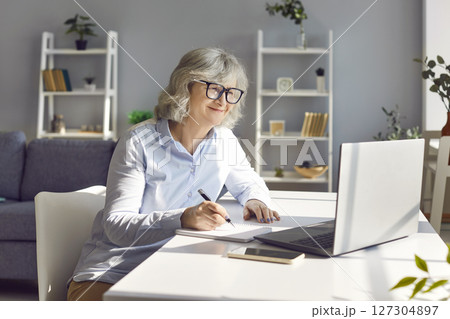 Senior smiling woman sitting on workplace at the desk working on laptop at home. Senior smiling woman sitting on workplace at the desk working on laptop at home. 127304897
