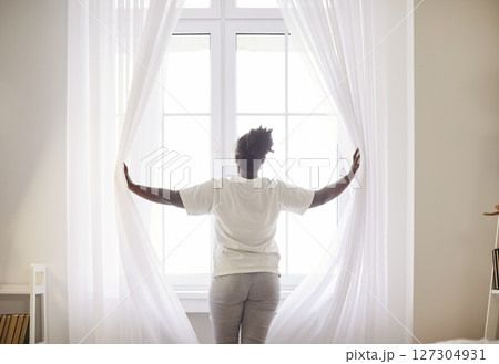 Young woman standing by the window opening curtains at home in the morning. 127304931