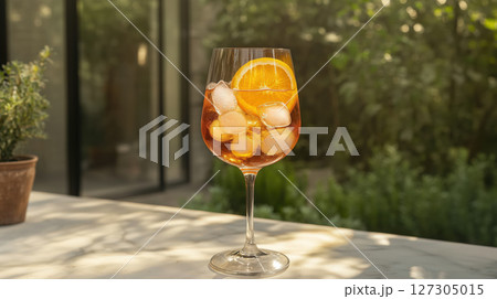 Aperol spritz cocktail in a wine glass with orange slice and ice cubes on a sunlit marble table outdoors 127305015