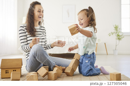 Joyful mother and daughter laughing together after knocking down wooden block tower during playtime. 127305046