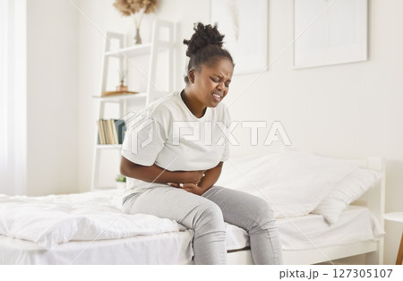 Sick african american woman sitting on bed putting hands on belly, suffering from strong bad stomach 127305107