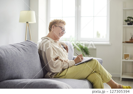 Mature woman sitting on sofa in living room at home and writing notes in her notepad. Mature woman sitting on sofa in living room at home and writing notes in her notepad. 127305394