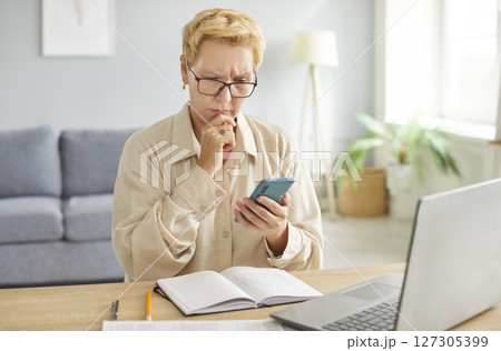 Pensive woman wearing glasses sitting at desk with laptop and phone, thinking about task or message. 127305399