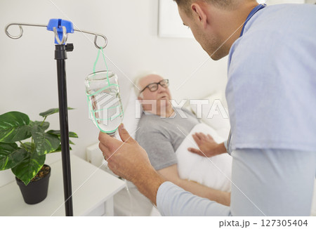 Caring young male nurse checking on elderly patient during IV infusion, providing support in clinic. 127305404