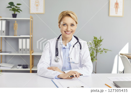 Portrait of cheerful young friendly female doctor in white medical uniform looking at camera. 127305435