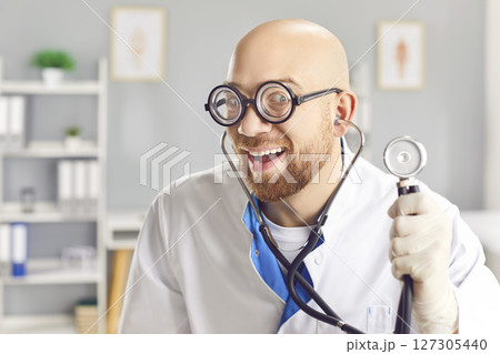 Portrait of a young funny smiling doctor in medical uniform standing with stethoscope in clinic. 127305440