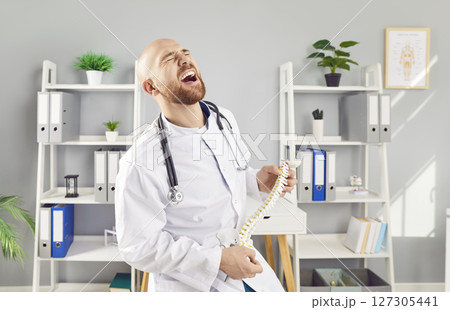 Funny young man doctor orthopedist playing on spine of human skeleton anatomical model in clinic. 127305441