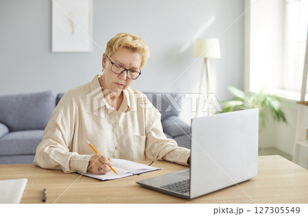 Senior busy mature woman wearing glasses writing notes, using laptop computer, sitting at table Senior busy mature woman wearing glasses writing notes, using laptop computer, sitting at table 127305549