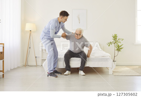 Portrait of a young friendly male nurse helping a senior patient to get off the bed at home. 127305602