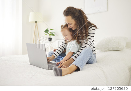 Smiling young mother and daughter sitting on big bed, using laptop, enjoying fun leisure together. 127305620