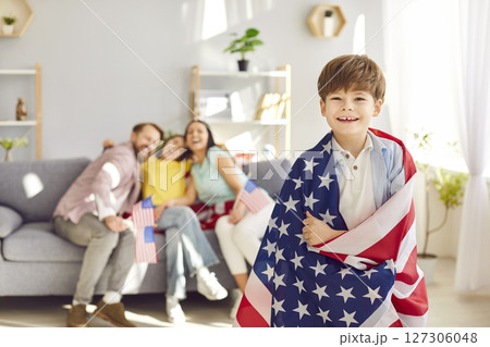 Child boy wrapped in us flag looking at camera with his happy american family in background. 127306048