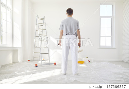 Male painter standing back in empty room holding a bucket and paint roller Male painter standing back in empty room holding a bucket and paint roller 127306237
