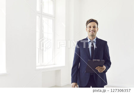Man realtor agent standing in empty apartments with documents in hands and looking at camera Man realtor agent standing in empty apartments with documents in hands and looking at camera 127306357