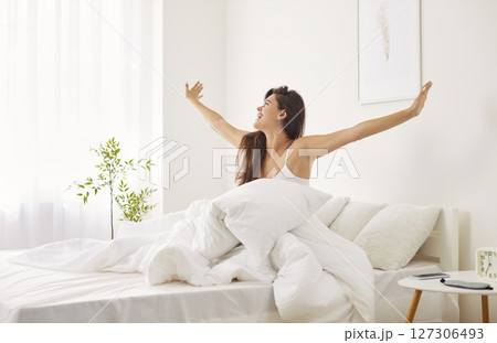 Happy Woman Waking Up And Stretching In Bed While Looking Out The Window 127306493