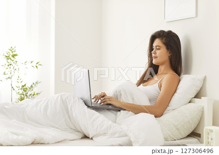 Relaxed young woman sitting in bed with laptop on knees, working from home or chatting with friends. Relaxed young woman sitting in bed with laptop on knees, working from home or chatting with friends. 127306496
