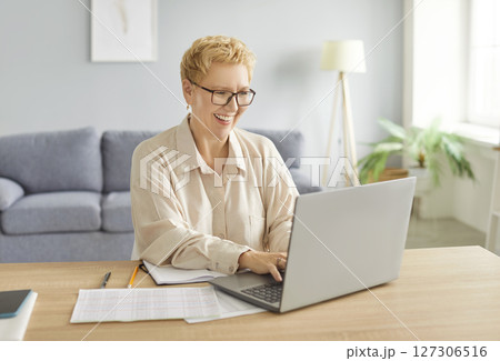 Happy smiling mature woman sitting on workplace at the desk working on laptop at home. 127306516