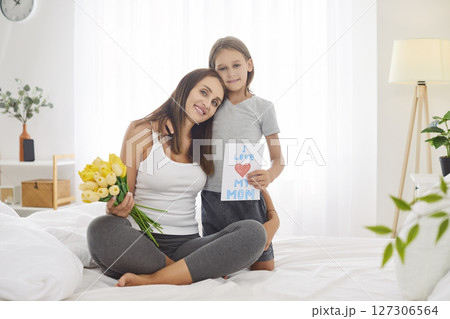 Child congratulating happy young mother with flowers bouquet and greeting card at home. Child congratulating happy young mother with flowers bouquet and greeting card at home. 127306564