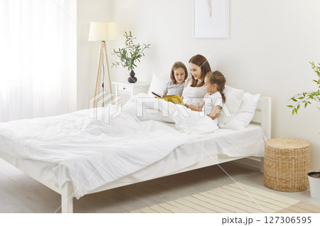 Young happy mother reading a book with her little cute children sitting on bed in bedroom at home 127306595