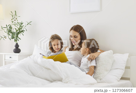Mother lies in bed with her two children in morning and reads book looking at illustrations. Mother lies in bed with her two children in morning and reads book looking at illustrations. 127306607