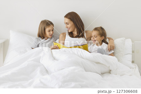 Smiling young mother sitting under cozy blanket with her children, reading fairytale book together. Smiling young mother sitting under cozy blanket with her children, reading fairytale book together. 127306608