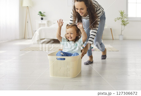 Young mother gives a laundry basket ride to her happy adorable little child daughter Young mother gives a laundry basket ride to her happy adorable little child daughter 127306672