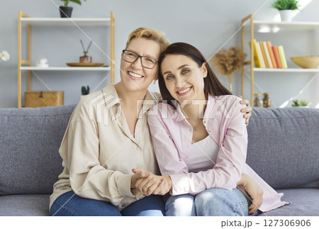 Happy adult daughter hugging her senior mature mother at home and looking cheerful at camera. Happy adult daughter hugging her senior mature mother at home and looking cheerful at camera. 127306906