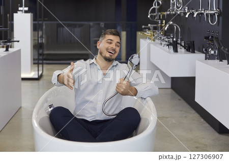 Cheerful man posing in bath tube in showroom holding shower head choosing bath supplies for bathroom Cheerful man posing in bath tube in showroom holding shower head choosing bath supplies for bathroom 127306907