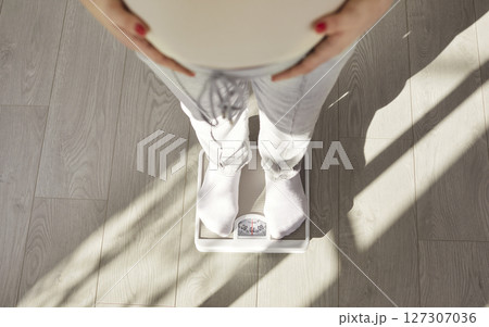 Cropped image of faceless pregnant woman standing on scales, controling her weight, touching belly Cropped image of faceless pregnant woman standing on scales, controling her weight, touching belly 127307036