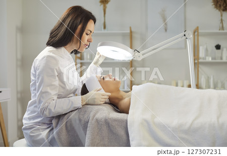 Professional female dermatologist cosmetologist, beautician doing female face exam study under lamp Professional female dermatologist cosmetologist, beautician doing female face exam study under lamp 127307231