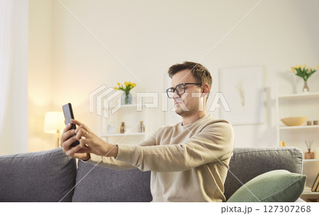 Focused man wearing glasses looking at phone screen from a distance, browsing mobile device apps Focused man wearing glasses looking at phone screen from a distance, browsing mobile device apps 127307268