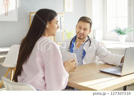 Smiling male doctor in white medical uniform meets to show checkup results on laptop, consult female 127307310