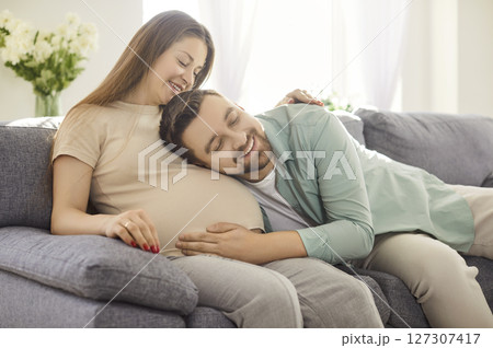 Happy man listening to his beautiful pregnant woman's tummy and smiling sitting on sofa at home. Happy man listening to his beautiful pregnant woman's tummy and smiling sitting on sofa at home. 127307417